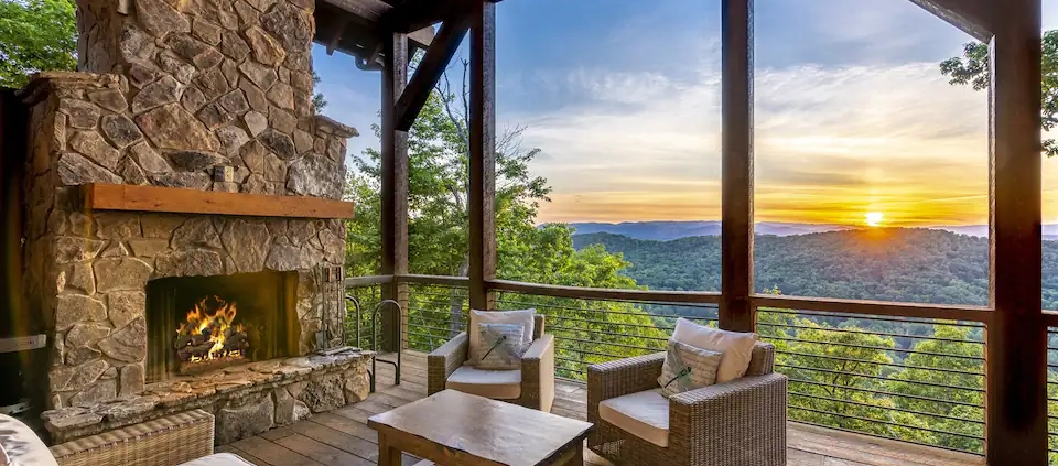 A mountain view of a cabin that has an outdoor porch with a fireplace and outdoor patio furniture, including a table, two chairs, and a day bed.